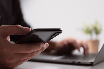 Aufkleber - Businessman hand using a smartphone and reading messages via an app while working on a laptop computer at a desk in the workplace.