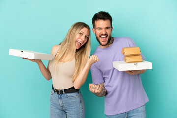 Wall Mural - Couple holding pizzas and burgers over isolated blue background celebrating a victory