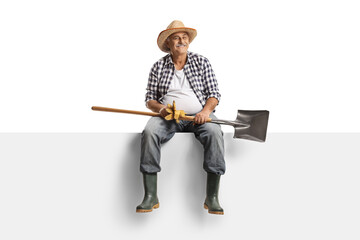 Cheerful mature farmer sitting on a blank panel, holding a shovel and smiling