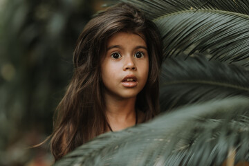 Wall Mural - The face of a little girl surrounded by tropical leaves. Closeup portrait of a beautiful swarthy baby with perfect skin and dark hair. Natural cosmetics, health, cleanliness, skin care, beauty concept