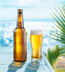 Wall Mural - Opened bottle of beer and beer glass with condensation on the wooden table. Blurred sparkling sea at the background. Summer vibes.