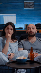 Wall Mural - Amazed couple watching movie on TV at night and eating popcorn, drinking beer discussing on film sequences . Focused concentrated astonished wife sitting home on confortable couch, facial expression
