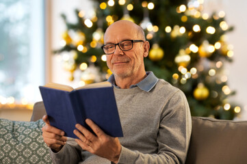 Sticker - winter holidays, leisure and people concept - happy bald senior man sitting on sofa and reading book at home in evening over christmas tree lights on background