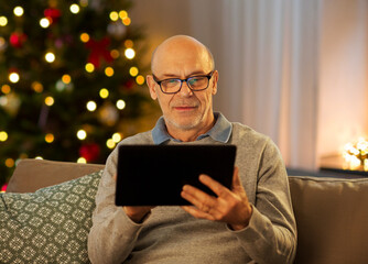 Wall Mural - technology, winter holidays and people concept - happy bald senior man with tablet pc computer sitting on sofa at home in evening over christmas tree lights background