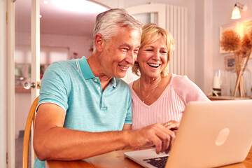 Wall Mural - Senior Retired Couple Sitting On Sofa At Home Shopping Or Booking Holiday On Laptop