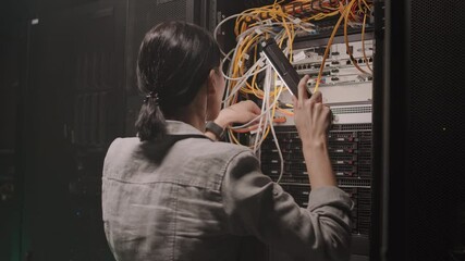 Rear medium close-up of dark-haired female Caucasian engineer holding flashlight, examining wire connection of server