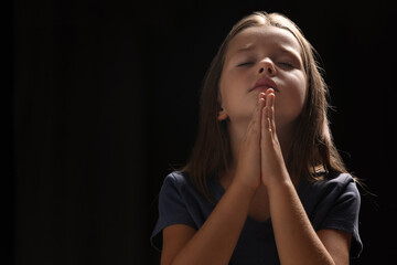 Cute little girl with hands clasped together praying on black background. Space for text