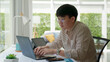 © ChayTee - Asia people happy man coding on laptop work remotely at home office sit relax smile write e-mail search data on desk. MBA online study class in reskill upskill college learning for job career path.
