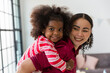Happy African American mother piggybacking with cute curly little daughter on back. Cheerful African mom playing, having fun and spending time together over Christmas tree at home. Merry Christmas