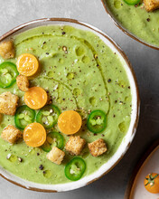 A Closeup View Of A Bowl Of Green Tomatillo Gazpacho Garnished With Jalapeno Slices, Cherry Tomatoes, Croutons, Olive Oil, And Ground Pepper