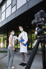 Canvas Print - asian reporter holding microphone near doctor in medical mask pointing away near blurred digital camera