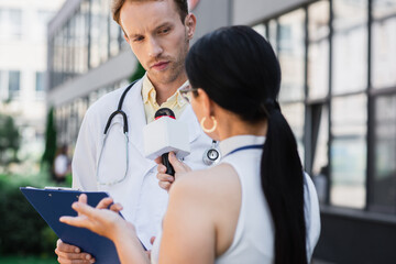 Wall Mural - doctor in white coat holding clipboard and looking at blurred journalist with microphone