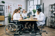 © sofiko14 - Aged male doctor with x ray scan in hands doing report to his international colleagues at office room. Multiracial practitioners cooperating for common medical research. One woman using wheelchair.