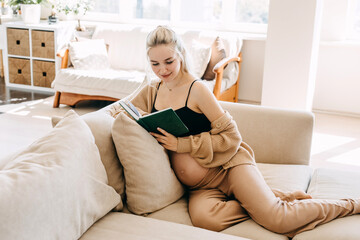 Wall Mural - Pregnant woman wearing cozy home wear, sitting on a sofa at home, reading a book.