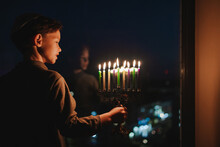 The Child Lights The Menorah For Hanukkah On The Windowsill. The Boy In The Kippah Sitting By The Window. Jewish Holiday. Tradition Is A Religious Ritual. Sunset. The First Star. Judaism
