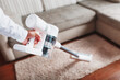 © Alexander - Powerful cordless vacuum cleaner with white cyclonic dust collection technology in hand, cleans the carpet in the house near the sofa.