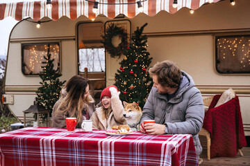 Celebrating Christmas in camper: young family drinking hot cocoa sitting outside Rv van decorated for Xmas celebration. Parents and daughter on road trip on winter holidays or vacation outside city