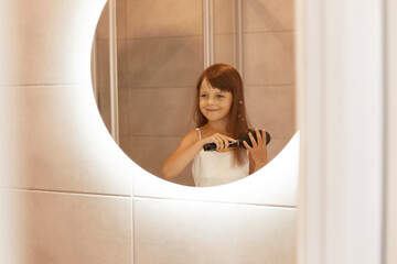 Wall Mural - Smiling little girl combing her hair in the bathroom in front of mirror, enjoying looking at her reflection, wearing home clothing, doing beauty procedures.