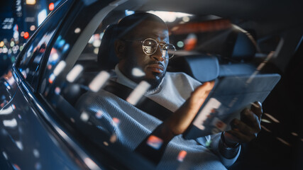 Wall Mural - Stylish Black Man in Glasses is Commuting Home in a Backseat of a Taxi at Night. Male Using Tablet Computer and Looking Out of Window while in a Car in Urban City Street with Working Neon Signs.