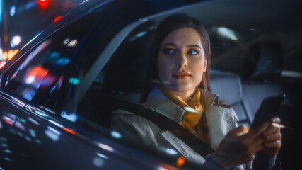 Wall Mural - Stylish Female is Commuting Home in a Backseat of a Taxi at Night. Beautiful Woman Passenger Using Smartphone and Looking Out of Window while in a Car in Urban City Street with Working Neon Signs.