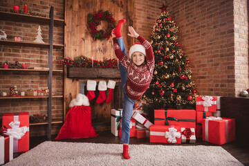 Photo of little kid sporty girl make stretch raise leg posing wear sweater santa hat socks in decorated room indoors