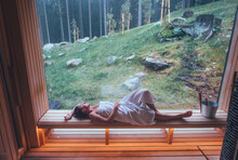 Relaxing Woman wrapped white towel lying on the wooden bench in Hot Finnish sauna with a huge wide window with green forest view and enjoying pleasant healthy body care temperature treatment.