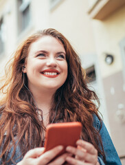 Wall Mural - Smiling red curly long hair caucasian teen girl walking on the street and browsing the internet using the modern smartphone. Modern people with technology devices concept image.