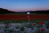 Fototapeta Kuchnia - Young woman poppy field. A brunette in a white hat among bright scarlet poppies poses against a purple sunset landscape. A beautiful wine red dress. The concept of freedom, romance, feelings, travel