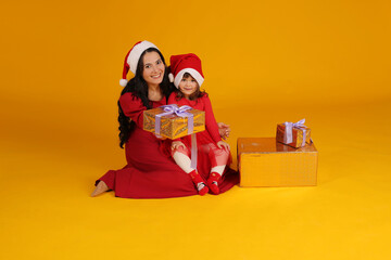 Wall Mural - beautiful brunette girl with long hair in a red dress santa claus hat next to a little daughter in a red dress santa claus hat gift box christmas concept