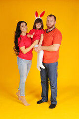 Wall Mural - a young family a brunette girl a guy with a red beard and a little daughter on her head rabbit pink ears in red T-shirts hugging yellow background