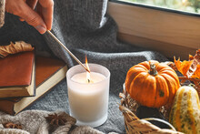 Hand With Burning Match Lighting A Candle On The Windowsill With Cozy Autumn Still Life With Pumpkins, Knitted Woolen Sweater And Books. Autumn Home Decor. Cozy Fall Mood. Thanksgiving. Halloween.