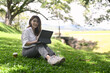 © Prathankarnpap - Happy woman sitting on grass with computer laptop in public park.