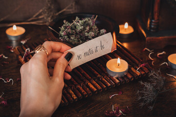 Wiccan witch holding a piece of paper with words So mote it be written on it, and burning it at her altar. Witch casting a spell