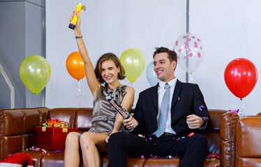 Caucasian businessman in business suit sitting on leather sofa with present gift box and balloons with female girlfriend in casual dress shoot paper confetti popper together celebrating xmas festival