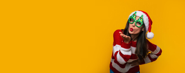 Wall Mural - Banner wide photo of Crazy and excited beautiful modern woman in funny Christmas glasses and Santa hat is having fun while posing against a yellow wall