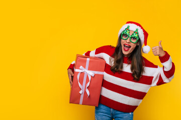 Wall Mural - Close up photo of beautiful excited smiling woman with Christmas gift box in hands is showing thumb up while posing on yellow background