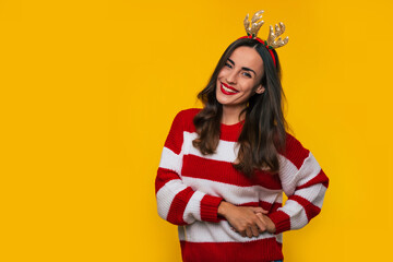 Wall Mural - Beautiful funny young woman in a striped winter sweater and deer hat on the head is posing isolated on yellow background in studio