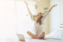 Beautiful Woman Sitting On The Bed In Front Of Her Laptop , Sprinkle Money On The Bed With A Happy Smile  Because She Profits From Investing In Online Stocks - Start Up Online Business Concept