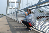Fototapeta Pomosty - Young beautiful fitness sporty jogger woman squats as taking a break on the bridge drinking water for hydration after cardio workout and training for healthy life by running outdoors