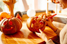 Close Up Of Woman Hands With Knife Carving Pumpkin. On The Table Lies A Orange Pumpkin With A Painted Horrible Face. Halloween, Decoration And Holidays Concept.