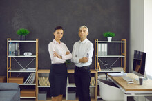 Serious Young Woman And Elderly Man In White Shirts Standing Arms Folded In Modern Office. Portrait Of Two Confident People, Business Professionals, Company Owners And Co Founders With Arms Crossed