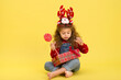© nellas - Little girl for the Christmas holidays with a gift and a lollipop on a yellow background to copy  space