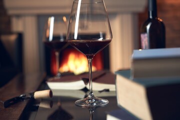 Two glasses of red wine with book and candle on table at home, fireplace in the background. Warm, dark colors.