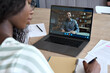 © insta_photos - Young black girl student having virtual remote class with distance indian teacher teaching training webinar or tutor on video call using laptop computer learning online at home. Over shoulder view