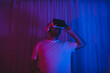© mckaya - Young man wearing virtual reality glasses in neon lit room.
