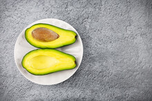 Ripe Avocado Cut Lengthwise Into Two Pieces On White Plate On Gray Surface