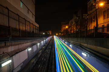 Colorful blue, yellow, green and red light trails from the CTA holiday train coming out from an underground tunnel at night with alley lights and residential buildings on either side of the tracks.