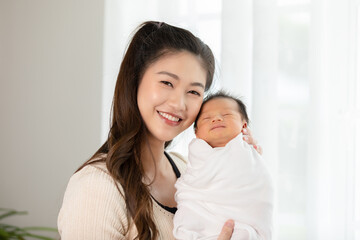 Beautiful Asian woman holding newborn baby in her arms standing in front of windows at cozy home.Happy infant baby sleep in mother arms safety and comfortable.Mom and Baby Concept