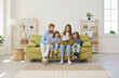 © Studio Romantic - Happy family reading book at home. Mom, dad, little kids sitting together on green sofa in new big light living room with white walls, beige brown rug, wicker storage basket, and shelves in background