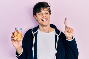 Handsome hipster young man holding salty biscuits smiling with an idea or question pointing finger with happy face, number one